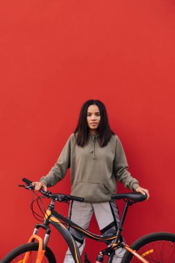 Kırmızı arka planda izole edilmiş bisikleti olan sıradan giyinmiş şık bir kadın mutlu bir yüzle kameraya bakıyor. Dikey.