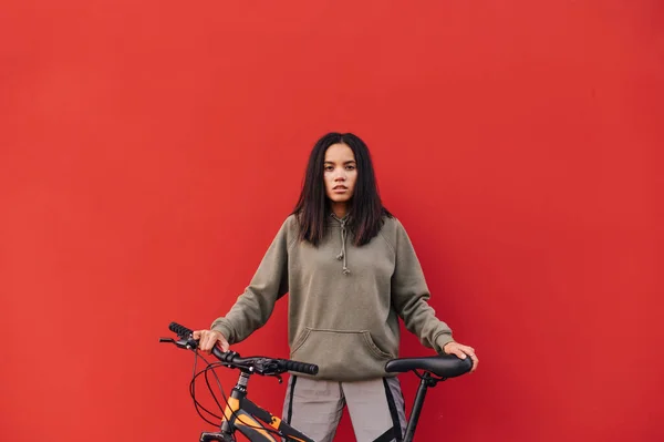 身穿运动服的漂亮女人站在一辆自行车的红色背景上 摆出严肃的姿势面对镜头 女模特在红墙的背景上摆着自行车的姿势 — 图库照片