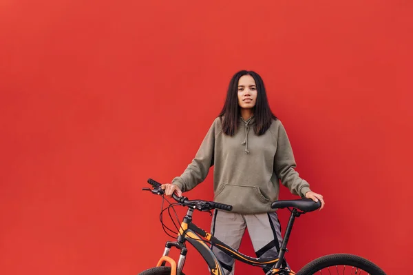 穿着时髦户外服装的积极的惊慌失措的女人站在那里 双手拿着一辆红色背景的自行车 面带微笑地看着摄像机 — 图库照片