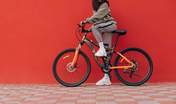 切り取られた写真 赤い壁を背景に自転車に乗った女性が通りに立っている サイクリング コピースペース 赤い背景の女性の足と自転車 — ストック写真