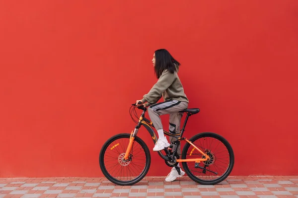 Beautiful woman rides a bicycle on a background of a red wall, looking ahead at the copy space. Hispanic woman standing on a bicycle on a red background.