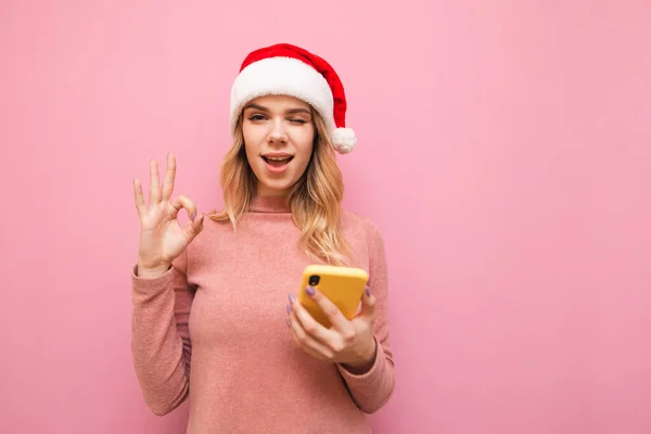 크리스마스 모자를 긍정적 소녀가 스마트폰을 배경에 카메라를 들여다보며 웃으며 괜찮은 — 스톡 사진