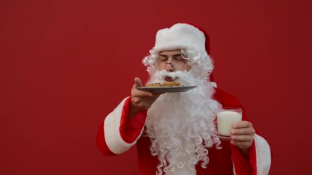 圣诞节有趣的圣诞老人一边站在红色的背景上一边闻巧克力饼干和牛奶 一边欣赏着它的芬芳 圣诞鸡窝 — 图库视频影像