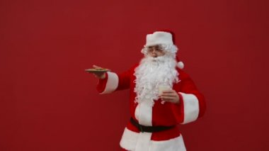 Kırmızı arka planda dans eden neşeli Noel Baba 'nın elinde bir tabak çikolatalı kurabiye ve bir bardak sütle. Noel konsepti.