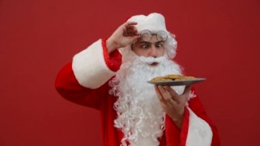 Elinde bir tabak kurabiyeyle şoke olmuş komik Noel Baba kırmızı arka planda duruyor ve komik bir yüzle plakaya ve kameraya bakıyor..