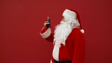Komik Noel Baba bir şişe kola açar ve mutlu yüzlü bir içki içer. Noel Baba bir şişe meşrubat açar ve kırmızı arka planda eğlenir..