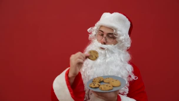 ポジティブなサンタは赤い壁の背景にプレートとおいしいチョコレートチップクッキーを食べます クリスマスのコンセプト — ストック動画