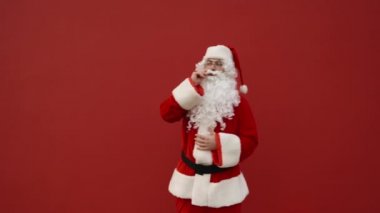Neşeli Noel Baba kırmızı arka planda duruyor, kameraya poz veriyor ve hafif bir el hareketi gösteriyor. Kırmızı duvarda dans eden Noel Baba.