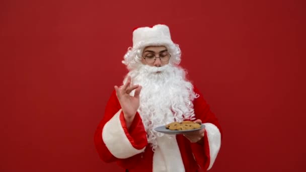 穿着圣塔服装的正人君子在红色背景下吃盘子里的圣诞饼干 红墙背景上挂着一盘巧克力饼干的快乐的桑塔 — 图库视频影像