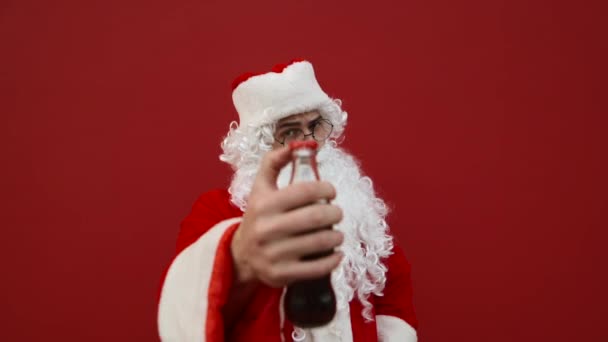 圣诞老人打开一瓶可乐 在红墙的背景下欢快地喝着一杯软饮料 穿着桑塔服装的男人在红色圣诞背景下喝可乐 — 图库视频影像