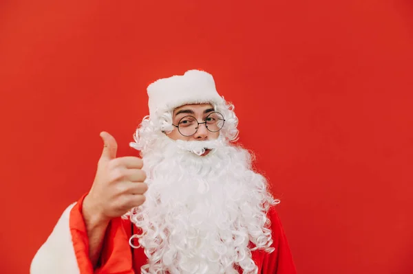 穿着圣塔服装的积极向上的男人 他脸上挂着笑容 竖起大拇指 在红色背景下被隔离的相机 新年及圣诞节 — 图库照片