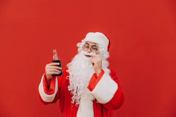 Santa Claus stands on a red background with a bottle of cola in his hands, twists his mustache and looks away at a sweet drink. Christmas cotnept.