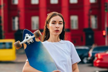 Şehrin arka planında elinde uzun bir sörf tahtası olan, kameraya dikkatlice bakan güzel bir bayan patencinin portresi..