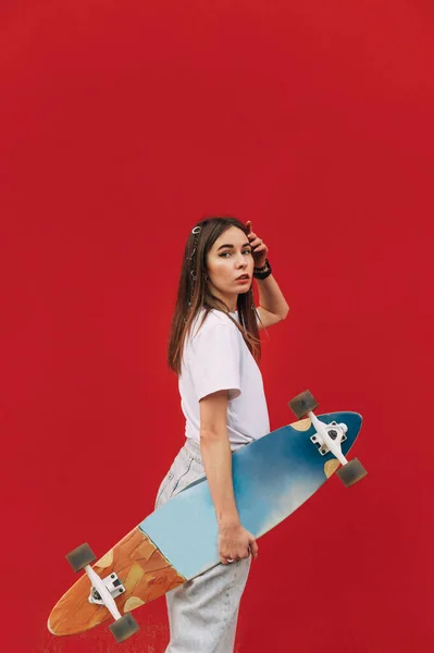 手にスケートで赤い背景にカメラをポーズしているカジュアルな服の美しい女性 スケートボードのコンセプト — ストック写真