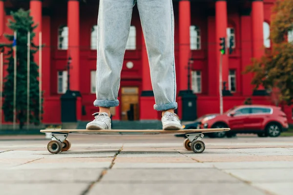 Zamknij Zdjęcie Kobiecych Nóg Dżinsach Tenisówkach Longboardzie Tle Czerwonej Architektury — Zdjęcie stockowe