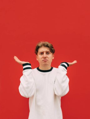 Kırmızı arka planda, şaşkın bir genç adamın portresi, üzgün bir yüzle kameraya bakıyor ve kollarını yana açıyor. Dikey.