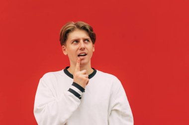 Komik düşünceli genç adam kırmızı arka planda duruyor, fotokopi odasına bakıyor ve düşünmek için bir jest yapıyor. Boşluğu kopyala