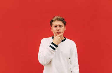 Ciddi bir genç adam kırmızı bir duvara yaslanır, beyaz bir kazak giyer, kameraya bakar ve düşünür, parmağını dudaklarına değdirir ve düşünceli bir şekilde bakar.