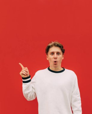 Kırmızı arka planda beyaz tişörtlü, kameraya bakan ve fotokopi alanında başparmağını gösteren şaşırmış genç adamın portresi. Dikey.