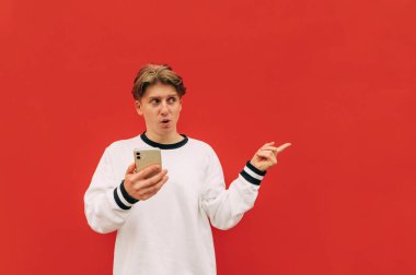 Elinde akıllı bir telefonla kırmızı arka planda duran ve yan tarafa bakıp fotokopi alanında parmağını gösteren şaşırmış bir gencin portresi.