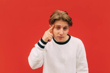Beyaz kazaklı genç bir adam kırmızı bir duvarın arka planında duruyor, parmağını şakağına doğrultuyor ve düşünceli bir yüzle kameraya bakıyor. Adam bir jest yapıyor.