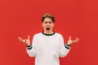 Kırmızı bir duvarın arka planında şaşırmış şeytani bir suratı olan kafası karışmış genç bir adamın portresi, kameraya çılgınca bakıyor ve elleriyle duygusal jestler yapıyor..