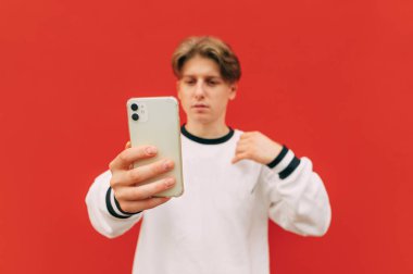 Beyaz giysili genç adam kırmızı arka planda duruyor ve akıllı telefon kullanıyor, akıllı telefona odaklan. Adam akıllı telefon kullanıyor, izole edilmiş..