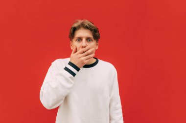 Şok olmuş genç adam elleriyle yüzünü kapatıyor ve kırmızı duvarın arka planında şok olmuş bir yüzle kameraya bakıyor. İzole edilmiş.