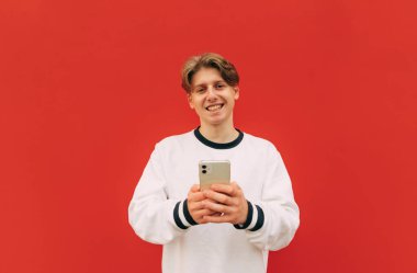 Parlak elbiseli adam elinde akıllı telefonla kırmızı bir duvarın arkasında duruyor, kameraya bakıyor ve gülümsüyor..