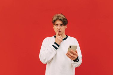Düşünceli genç bir adam elinde akıllı bir telefonla kırmızı bir arkaplanda duruyor ve özenle fotokopi alanının kenarına bakıyor. Kırmızı duvar arkaplanında izole.