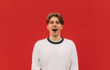 Kırmızı arka planda izole edilmiş beyaz kazaklı, şaşkın yüzüyle kameraya bakan, şaşırmış, pozitif genç bir adam..