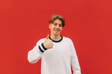 Kırmızı arka planda duran mutlu genç adam ve kırmızı arka planda baş parmak işareti yapıyor, kameraya bakıyor ve gülümsüyor.