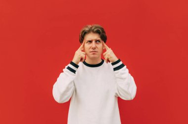 Beyaz bluzlu dalgın bir adam kırmızı bir duvarın arka planında duruyor ve üzgün bir yüzle parmaklarını şakağına gösteriyor. Tecrit edilmiş bir adamda düşünceler baş ağrısı.
