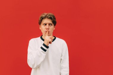 Sıradan giyinmiş komik bir adam kırmızı arka planda duruyor, fotokopi odasına bakıyor ve sessizlik işareti yapıyor. Genç adam parmak hareketini gösteriyor. İzole edilmiş.