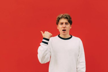 Beyaz kazaklı genç adam kırmızı arka planda duruyor, kameraya bakıyor ve boş bir noktaya parmağını doğrultuyor. İzole edilmiş