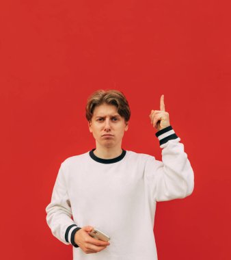 Sıradan kıyafetler giymiş, elinde akıllı telefonuyla üzgün bir genç kameraya bakar ve fotokopi uzayında başparmağını gösterir. Kırmızı duvar arkaplanında izole.