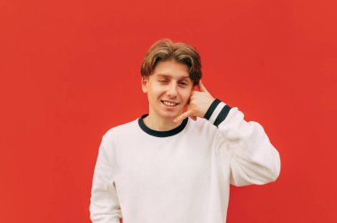 Beyaz kazaklı neşeli bir adam yüzünde bir gülümsemeyle kameraya bakar ve kırmızı duvar arkasında izole edilmiş bir çağrı hareketi gösterir..