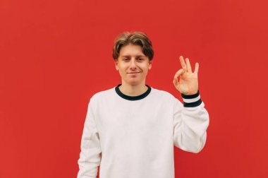 Pozitif adam parmaklarıyla iyi bir hareket gösteriyor ve kırmızı duvarda mutlu bir yüzle kameraya bakıyor. Genç adam jestini gösteriyor ve gülüyor..