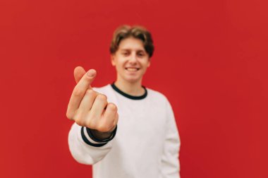 Kalp jestinin yakın fotoğrafı. Geçmişi. Yüzünde gülümseme olan sevimli bir adam parmaklarını kırmızı arka planda bir kalp jesti olarak gösteriyor, mutlu bir yüzle kameraya bakıyor..