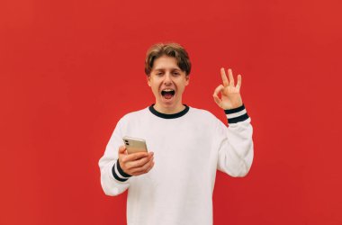 Elinde akıllı telefon olan mutlu genç adam kameraya bakar ve parmaklarıyla iyi bir hareket gösterir. Kırmızı arkaplanda izole.