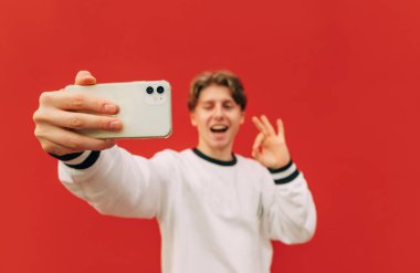 Hafif günlük kıyafetlerle gülümseyen adam, kırmızı arka planda akıllı telefondan özçekim yapıyor, yüzünde gülümseme ve iyi bir jest gösteriyor. Akıllı telefona odaklan..