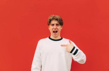 Beyaz kazaklı duygusal genç adam kendini işaret ediyor ve kameraya hoşnutsuz bakıyor. Tatminsiz adam kendini kırmızı bir duvarın arka planına işaret ediyor.