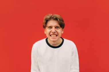 Kırmızı bir duvarın arka planında sıradan giyinmiş kötü bir çocuğun portresi, kameraya bakıyor ve kızgın. İzole edilmiş. Kızgın genç adam kırmızıda izole edilmiş..