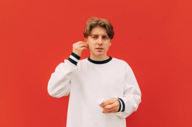 Yakışıklı genç adam şık bir yumurta geyiği kıyafeti içinde kırmızı arka planda duruyor ve müzik dinlemeden önce kulaklığına kablosuz kulaklık takıyor. İzole edilmiş.