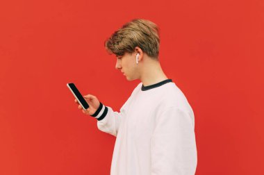 Kablosuz kulaklıklı adam kırmızı bir duvarın arka planında akıllı telefon kullanıyor, yüzünü çeviriyor ve ekrana bakıyor. Genç bir adam yürürken akıllı telefon kullanıyor..