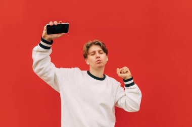 Şık giyinmiş neşeli bir adam kulaklıkla müzik dinliyor ve kırmızı arka planda gözleri kapalı, elinde akıllı bir telefonla dans ediyor..