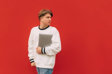 Kablosuz kulaklıklar takmış, elinde dizüstü bilgisayar olan yakışıklı bir adam kırmızı duvarın arka planında yürür ve boş bir yere bakar.