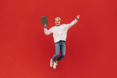 Mutlu genç adam elinde bir dizüstü bilgisayarla kırmızı arka planda zıplıyor ve yüzünde bir gülümsemeyle kameraya bakıyor. İzole edilmiş.