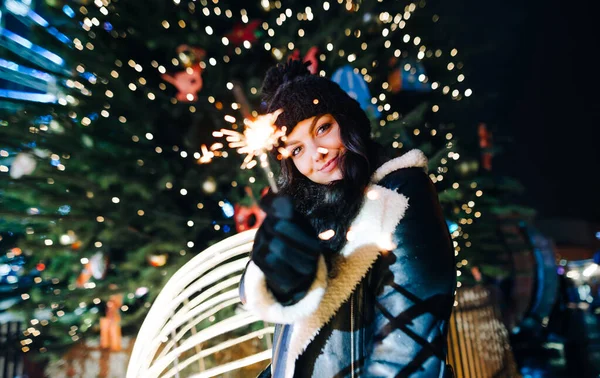 クリスマスツリーの背景にキラキラと輝く手でクリスマスの街に立つ魅力的な女の子は かわいい笑顔でカメラを見ています 新年とクリスマスのお祝い — ストック写真