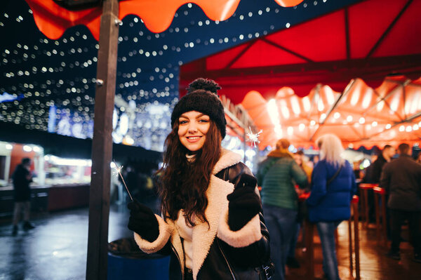 Happy girl posing for the camera with lighted sparklers in her hands, looking at the camera and smiling. Lady lights sparklers in the evening at the evening Christmas market.